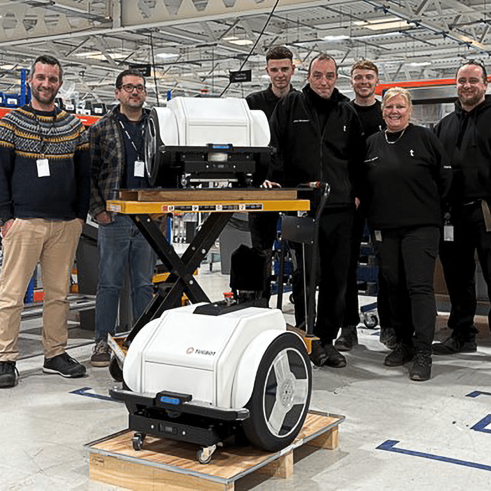 Tharsus and Tugbot engineers standing behind two Tugbot 2 autonomous mobile robots on the factory floor, celebrating a production milestone.